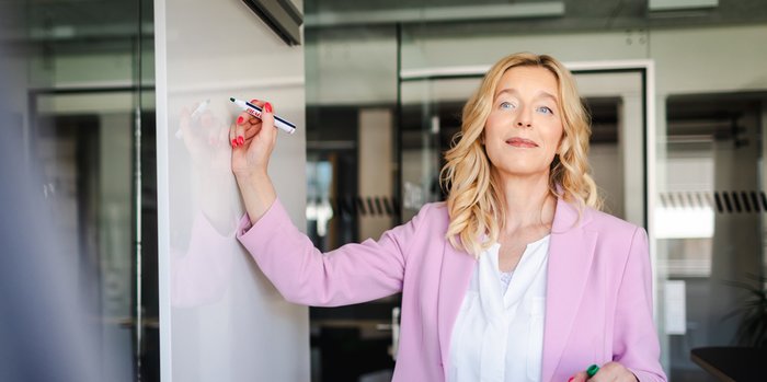 Persönlichkeitsentwicklung Beratung von Unternehmensberaterin Beatrix Becker – sie steht in einem modernen Büro, trägt ein rosa Blazer und schreibt mit einem Marker auf ein Whiteboard.